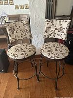 Front view of two swivel stools with beige fabric upholstery featuring brown leaf designs, metal frames, and wide cushioned seats and backs.