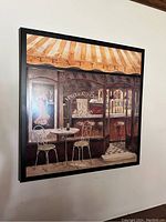 Photo of square wall canvas hanging on white wall showing an Italian trattoria storefront with chairs and table outside.