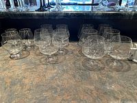 Photo showing a total of 14 assorted whiskey snifters arranged on a countertop. Different glasses with varying textured vertical ribbing and smooth glass bowls.