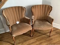 Photo of two antique armchairs with brown velvet upholstery and wooden curved frames positioned on wooden floor against a white wall.