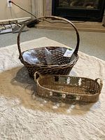 Two wicker baskets placed on carpet floor. One is a dark brown round basket with a handle, the other a light natural rectangular basket with painted accents and side handles.