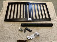 Black wooden baby gate with vertical bars laid flat, hardware components below it including screws and cylindrical piece.