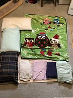 Photo showing six fitted sheets in various colors, three pillows, and one owl-themed fleece blanket arranged on carpet