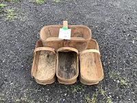 Photo showing four wooden berry baskets arranged on gravel ground. Each basket is rectangular with rounded corners and has a wooden slatted base reinforced by wooden side bands. The baskets show signs of use with discoloration and some surface wear.