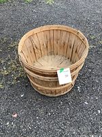 Photo of a round wooden bushel basket with vertical slats and horizontal bands, showing dirt and wear marks, placed on gravel ground.