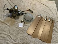 The ceiling fan is disassembled into parts: the main metal housing with a frosted glass light cover, four wooden blades, and mounting hardware. The photo shows the fan laid out on a fabric surface.