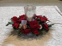 Front view of the Christmas centerpiece showing artificial red poinsettias surrounding a cylindrical mercury glass candle holder, placed on a light-colored textured fabric.