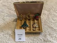 Five Christmas-themed pins in a gold-colored case with velvet-like insert, shown on a carpeted surface.