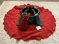 Photo showing red fabric tree skirt spread on floor, green plastic tree stand placed on top, and red angel tree topper standing in front.