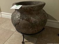 Side view of the large clay pot showing grapevine designs and part of the metal stand.