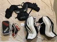 Photo of various pieces of youth goalie equipment laid on beige surface including leg pads, chest protector, blocker, and a pair of gloves.