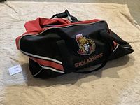 Photo showing side of a red and black hockey bag featuring the Ottawa Senators logo and wordmark, displayed on a beige surface.