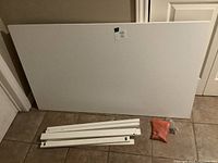 Dismantled IKEA MELLTORP table components including white rectangular tabletop and four white legs placed on tiled floor.