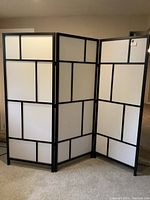 Full view of the three-panel IKEA RISÖR room divider showing white opaque panels framed by black sections with geometric pattern and floor placement.