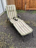 Lounger chair with striped cushions and black solar light sitting on it outdoors on gravel and grass with wooden fence in background.