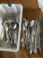 White plastic container with mixed forks, spoons, and knives inside, seen from above with lid beside.