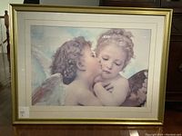 Photo of the framed print showing two cherubic children, one kissing the other, with gold frame and off-white matting.