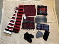 Photo showing five scarves arranged in two rows with patterns including stripes and plaids in colors red, black, grey, blue, and maroon. One long striped hand knit scarf is placed to the left. Below the scarves are two black hand knit slippers, one grey knit hat, and one pair of black suede gloves.