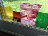 Three stained glass panels on a windowsill with grass outside. The colors are amber yellow, deep orange, marbled red, and marbled green.