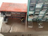 Photo showing two plastic organization boxes with multiple small drawers containing various hardware items, placed on a cardboard surface, dusty and soiled.
