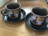 Photo of two brown ceramic owl mugs each paired with a matching dark saucer set on a wooden surface.