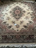 Full view of the large wool rug with intricate floral and medallion designs on beige background with fringed edges.