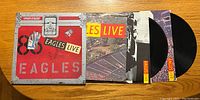Two black vinyl records partially pulled out of album cover with Eagles Live design in red and silver.
