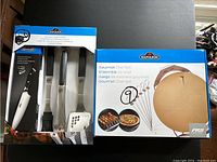 Front image showing boxed 4-piece BBQ toolset with knife, brush, tongs, and spatula alongside boxed round pizza stone