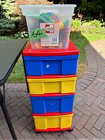 Clear plastic bin with assorted large Lego blocks on top of multi-colored plastic storage cart outdoors.