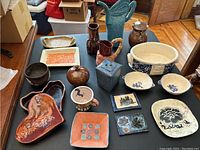 Wide view of assorted pottery items on black tabletop, including pitchers, vases, bowls, plates, and trays with diverse shapes and glazes.