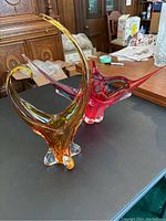 Two freeform glass sculptures, one red and one amber gradient, displayed side by side on a black surface with room background