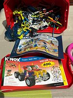 Bin of assorted K'NEX plastic construction parts spilling out, including rods, wheels, and mechanical components with two K'NEX printed booklets lying in front.