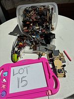 Photo showing assorted LEGO pieces spilling out of a plastic bin, with a pink drawing tablet marked 'LOT 15' in the foreground