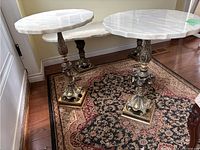 Pair of round side tables with white marble tops and decorative brass bases shown on rug in front of door with wall.