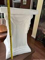 Side view of white plaster plant stand showing fluted vertical grooves and garland motif with measuring tape indicating height about 21 inches