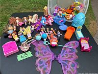Photo shows large assortment of small dolls and toys on black surface with hand-labeled number 65. Includes several Polly Pocket dolls, plastic furniture pieces such as a small chair, drawer units, beds, and a set of large glitter fairy wings.