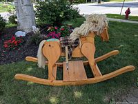 Side angle showing solid wood rocking horse with yarn mane and tail, adjustable stirrups with black and white checkered saddle, and wooden curved rockers on grass.