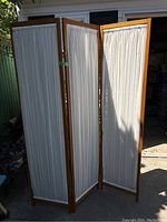 Full view of the 3-panel wooden room divider with white pleated cloth inserts, front angle with outdoor background.