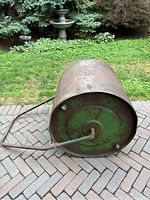 Photo of rusty green steel lawn roller lying on its side on a brick patio with a garden background, showing the cylindrical shape and round end with handle attached.