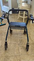 Front view of the blue metal walker showing the seat, wheels, and padded horizontal support bar.