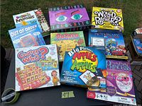 Photo showing a collection of various board and card games arranged on grass including Guess Who?, Sudoku, Apples to Apples, Pictionary, Pretty Pretty Princess, and others.