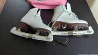 Pair of white woman's figure skates size 8.5 on a dark surface with pink and grey backpack visible in background, small skate blade guard and tool alongside.