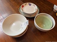 Overview of all bowls stacked on wooden surface showing various bowl types including plain white ribbed bowls, vegetable motif rim bowls, and green pottery bowl.