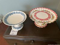 Two ceramic cake stands on a wooden surface, one with blue grapevine relief (Wedgwood), the other with pink floral pattern (Crown Staffordshire).