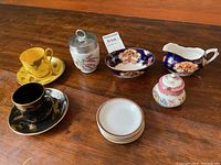 Full lot shown including 2 cups and saucers (yellow and black), lidded sugar bowl, small stacked plates, Royal Albert creamer, Royal Worcester egg coddler, and floral blue sugar bowl with handle.
