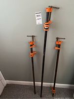 Three heavy metal pipe clamps standing upright against wall, showing varying lengths and orange clamp screws.
