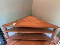Wooden triangular corner table with two lower shelves, placed in a corner.