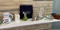 Full view of the lot items on shelf including Lampe Berger lamp, ceramic pitchers, glass pitcher, shakers, glass baskets, ashtray