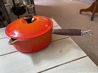View of Le Creuset red enamel cast iron saucepan with lid, wood handle, and spout showing some wear and chips in the finish.