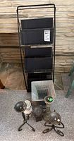 Photo showing black metal three-tier magazine rack and various candle holders including metal, wood, and ceramic, plus square metal urn on floor beside it